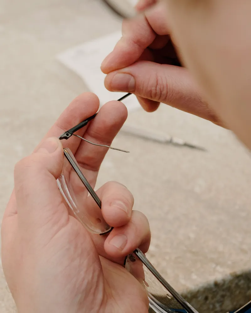 Manos trabajando en la mesa del taller mientras fijan la varilla al frontal de unas gafas con un destornillador fino
